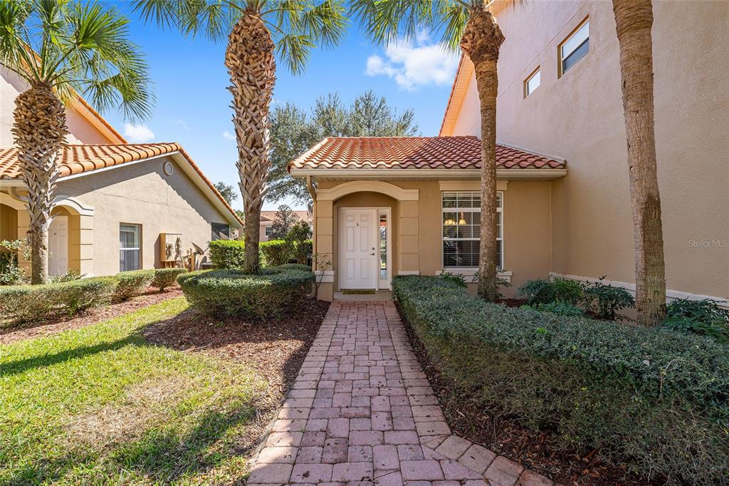 A brick walkway leads to the front entrance of a tan house with palm trees — 5427 COMPASS POINT UNIT 1, OXFORD, FL