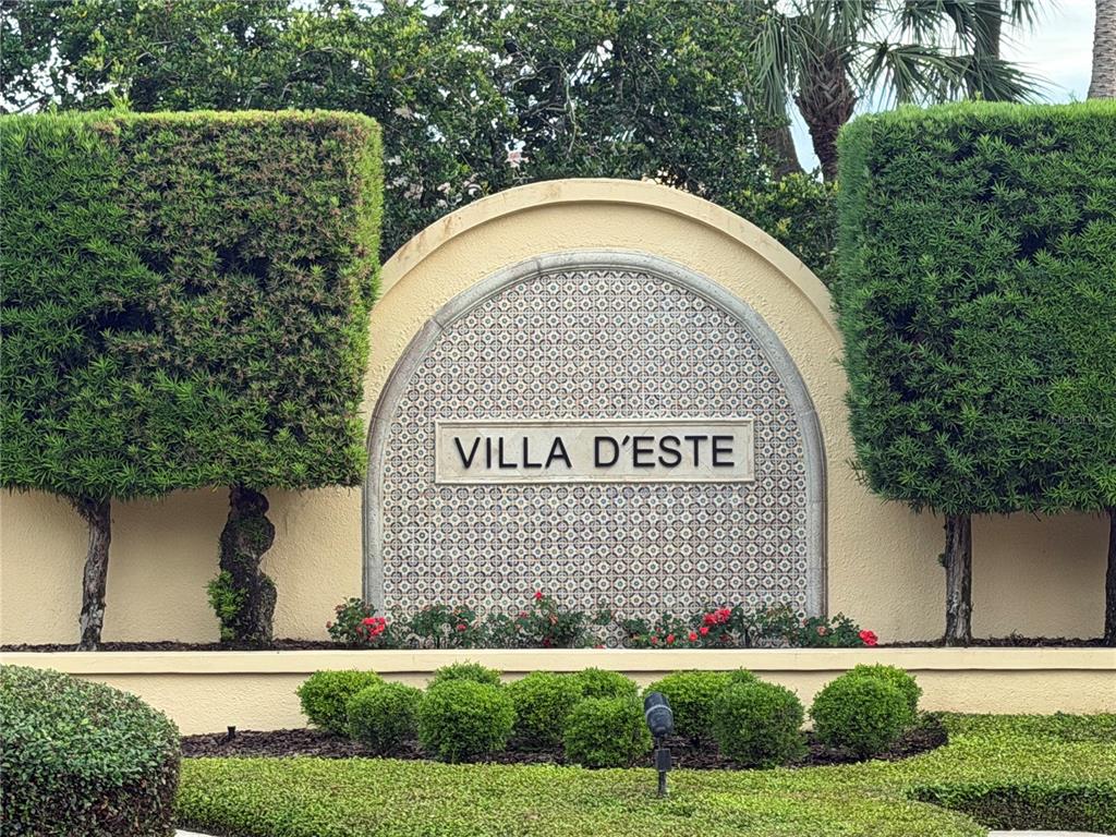 A sign reading Villa D'Este is centered on a stucco wall between two manicured trees — 2428 VIA GENOVA, APOPKA, FL