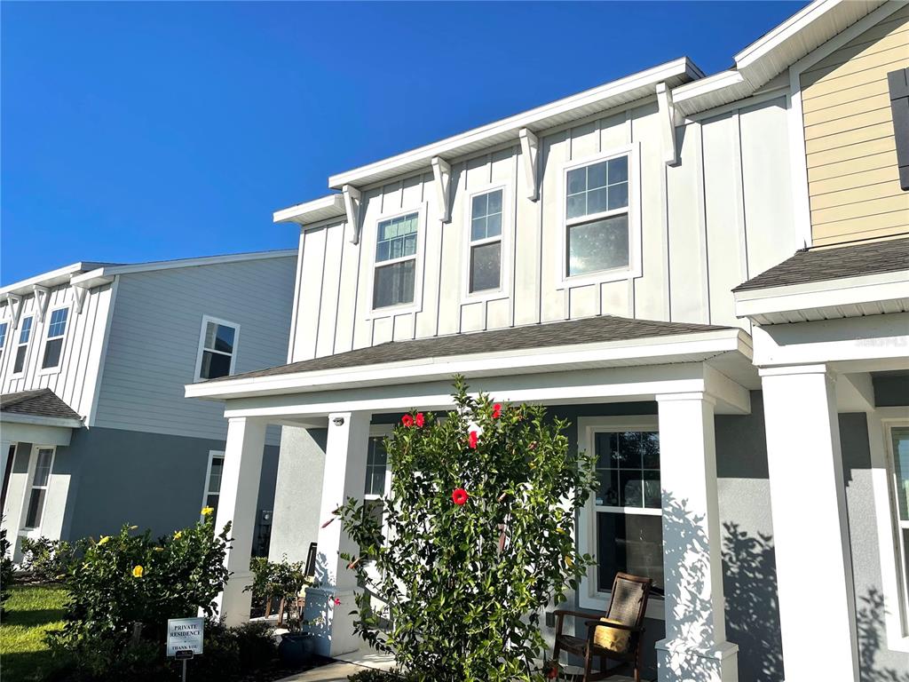 A two-story house exterior features white board-and-batten siding, a covered porch, and red hibiscus flowers — 5211 AVALON PARK BOULEVARD, WESLEY CHAPEL, FL