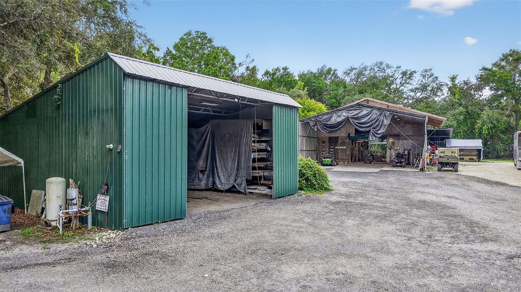 A green metal storage shed stands next to a larger open-air workshop on a gravel lot — 1862 S US 301, SUMTERVILLE, FL
