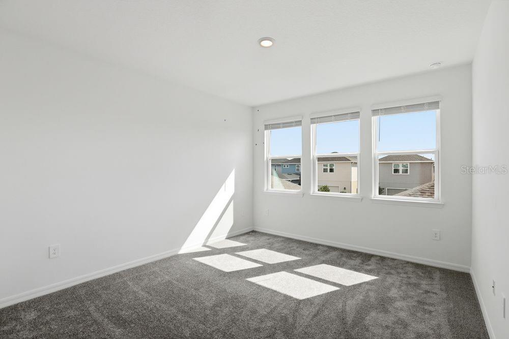 An empty bedroom features gray carpet, white walls, and three windows overlooking neighboring houses — 16050 HONEY HARVEST STREET, WINTER GARDEN, FL