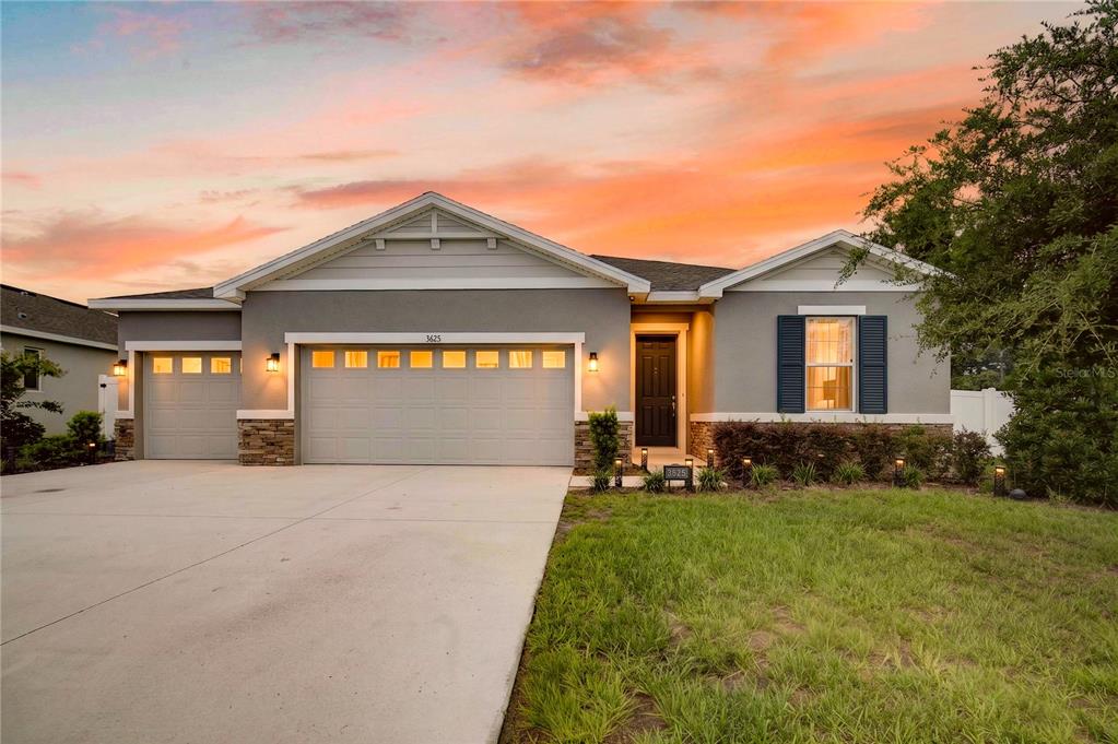 A single-story suburban house features a three-car garage, gray stucco siding, and a sunset sky — 3625 SUBLETT LOOP, TAVARES, FL