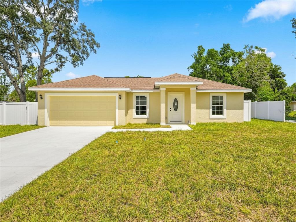 A single-story yellow house with a two-car garage and white vinyl fencing under a blue sky — 7209 APACHE TRAIL, SPRING HILL, FL
