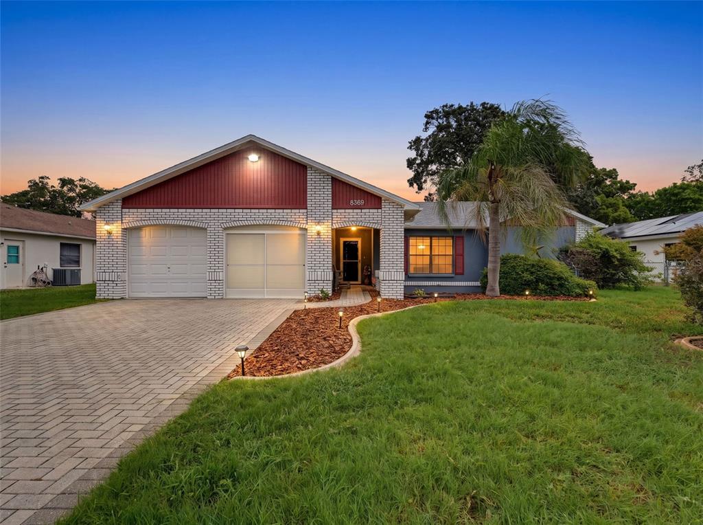 A single-story house with a brick facade, red gable, and a paved driveway at dusk — 8389 CAMPHOR DRIVE, SPRING HILL, FL