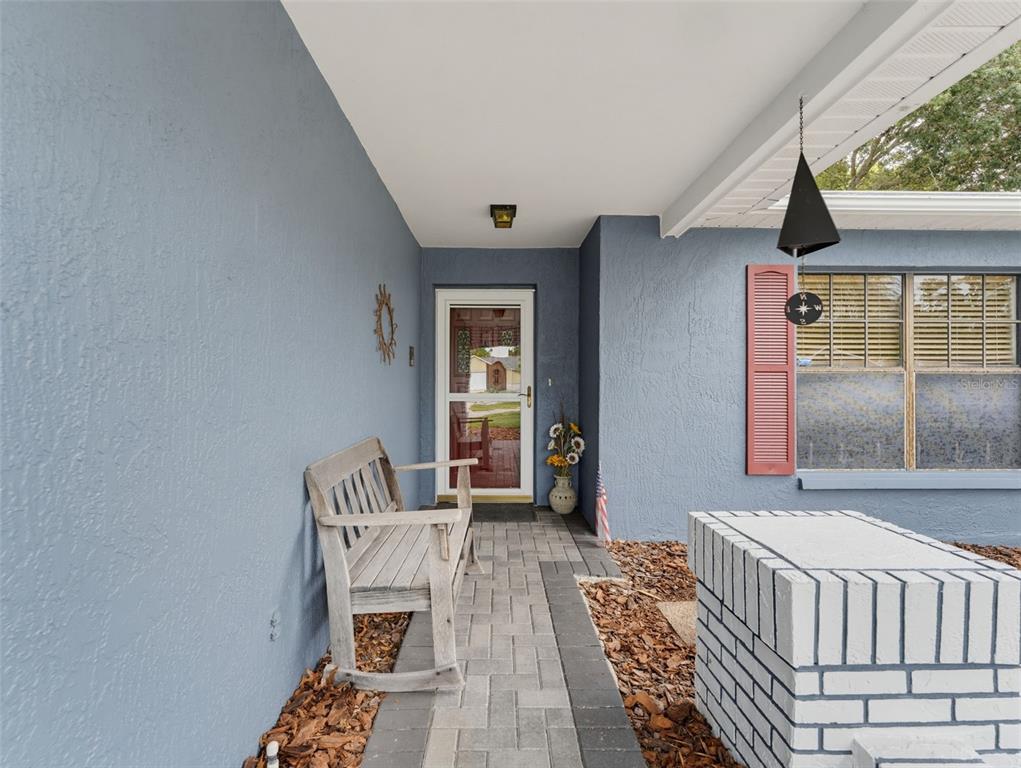 A covered porch features a paver walkway, a wooden rocking bench, and a brick planter — 8389 CAMPHOR DRIVE, SPRING HILL, FL