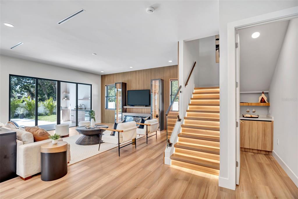 A modern living room features a slatted wood accent wall, illuminated staircase, and glass sliding doors — 2112 W FARWELL DRIVE, TAMPA, FL