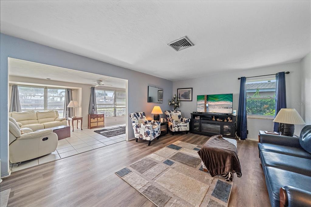 A living room with wood-look flooring, two patterned armchairs, a television, and an adjacent sunroom — 3286 BRUNSWICK LANE UNIT 704, SARASOTA, FL