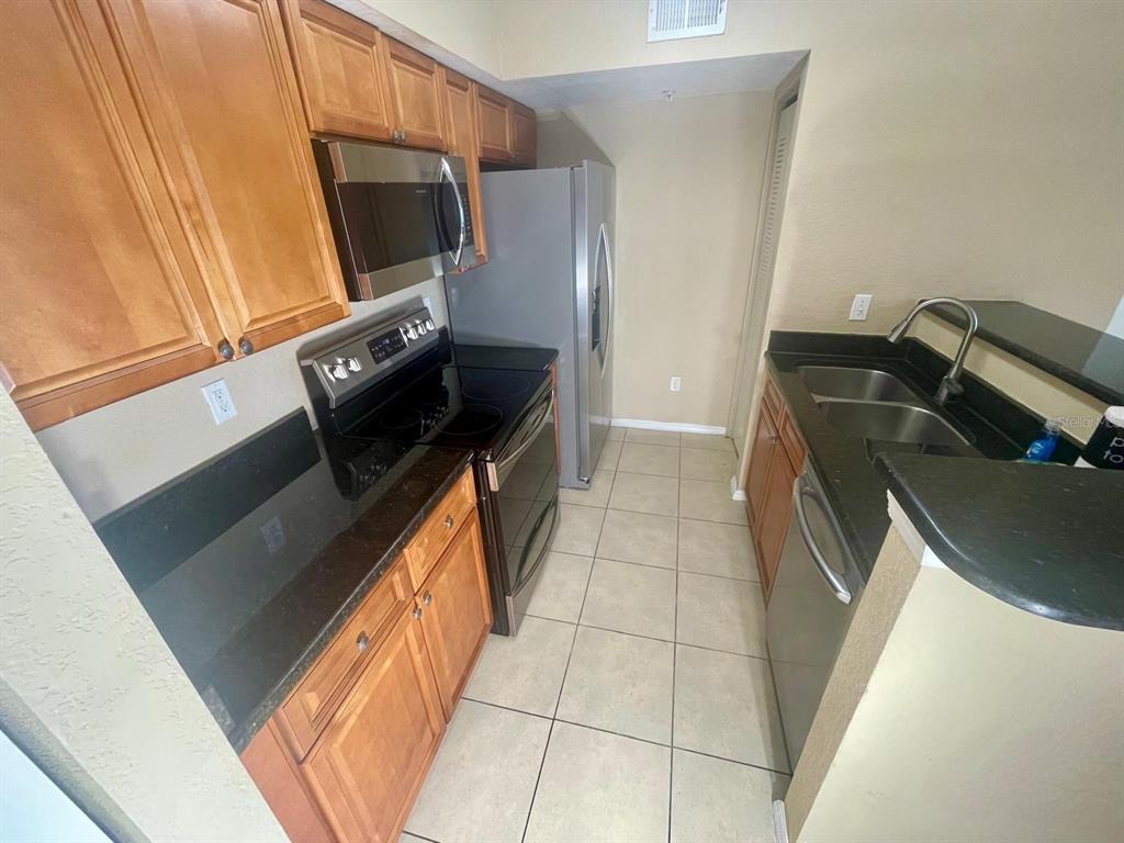 A kitchen features wood cabinets, black countertops, stainless steel appliances, and light-colored tile flooring — 4103 TROPICAL ISLE BOULEVARD UNIT 138, KISSIMMEE, FL