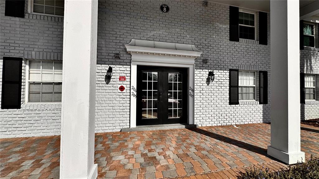 The exterior entrance of a white brick building features double glass doors and a paved patio — 205 LEWFIELD CIRCLE UNIT 205, WINTER PARK, FL