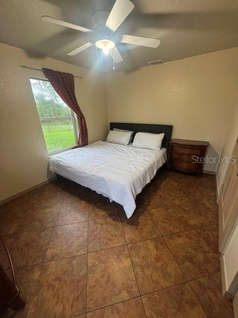 A bedroom features a bed with white linens, a ceiling fan, and brown tile flooring — 14373 IRVING STREET, SPRING HILL, FL