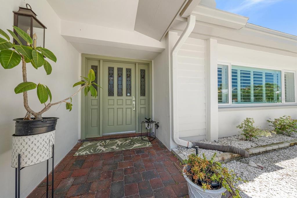 A sage green front door with sidelights sits on a brick porch near a potted plant — 821 JACARANDA DRIVE, LARGO, FL