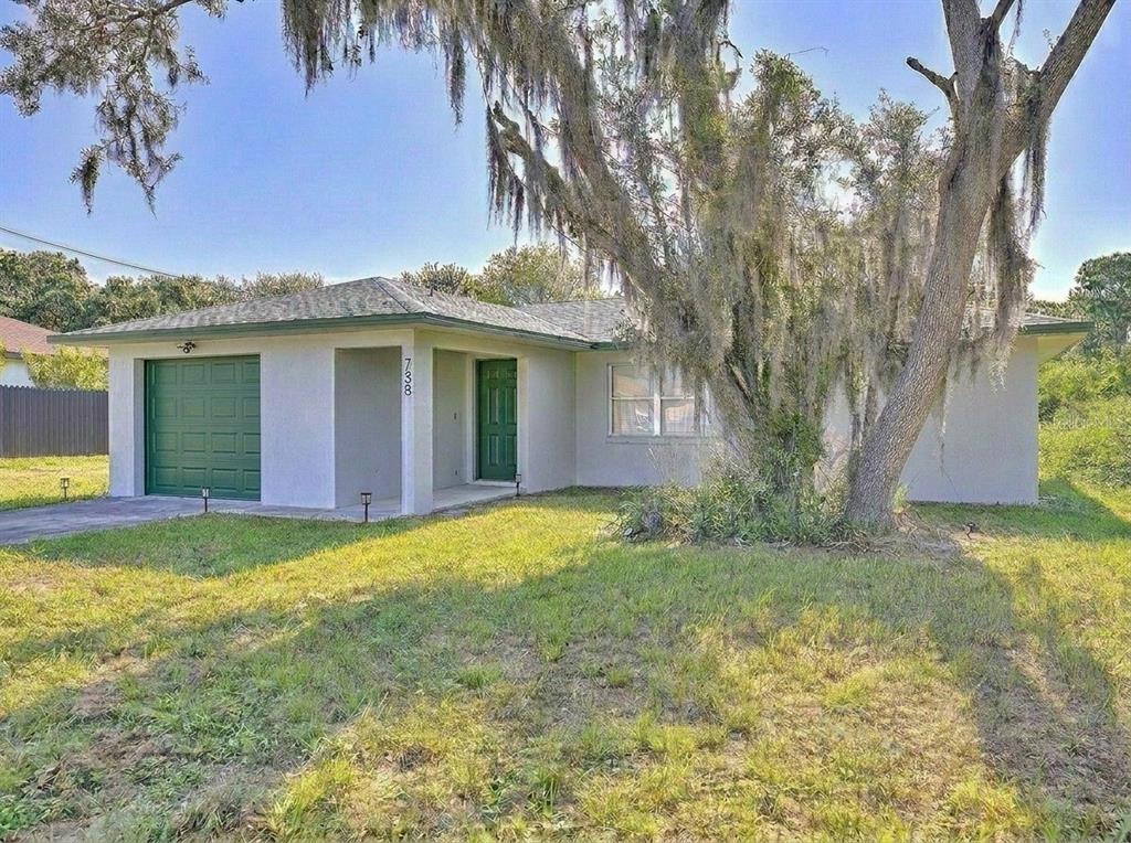 A single-story house with a green garage door and front door sits behind a large tree — 738 LEMANS DRIVE, SEBRING, FL