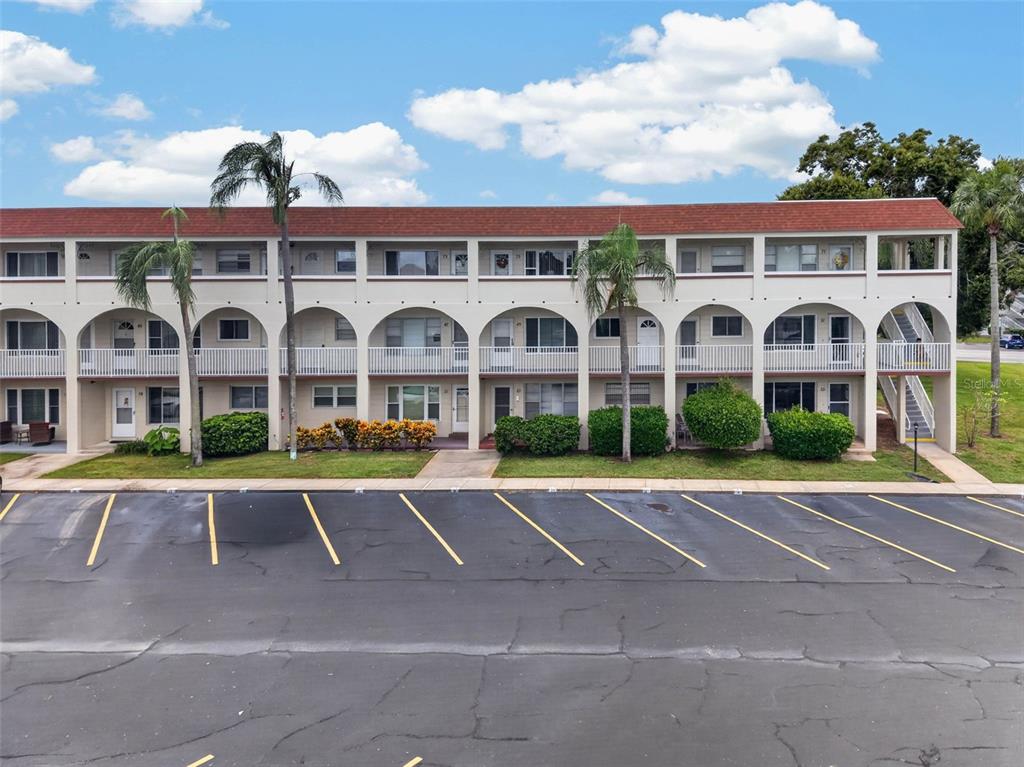 A three-story apartment building features arched balconies, a red roof, and a paved parking lot — 2433 BRAZILIA DRIVE UNIT 49, CLEARWATER, FL