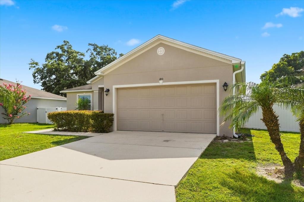A single-story house exterior features a beige stucco facade, a two-car garage, and a driveway — 180 SOLIS DRIVE, WINTER HAVEN, FL