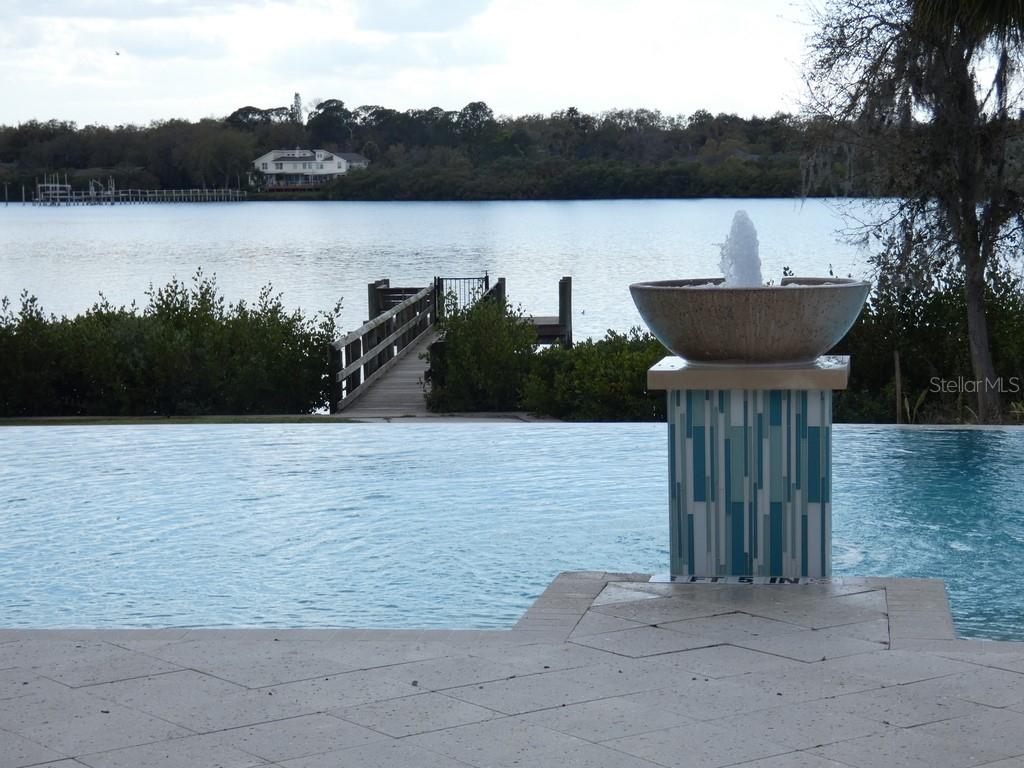 A fountain sits on a tiled pedestal beside an infinity pool overlooking a lake — 1505 SUNSET WIND LOOP, OLDSMAR, FL
