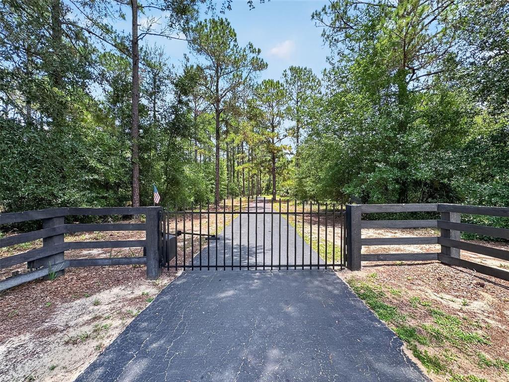 A black metal gate spans a paved driveway flanked by wooden fencing and dense trees — 22625 LAKE SENECA ROAD, EUSTIS, FL