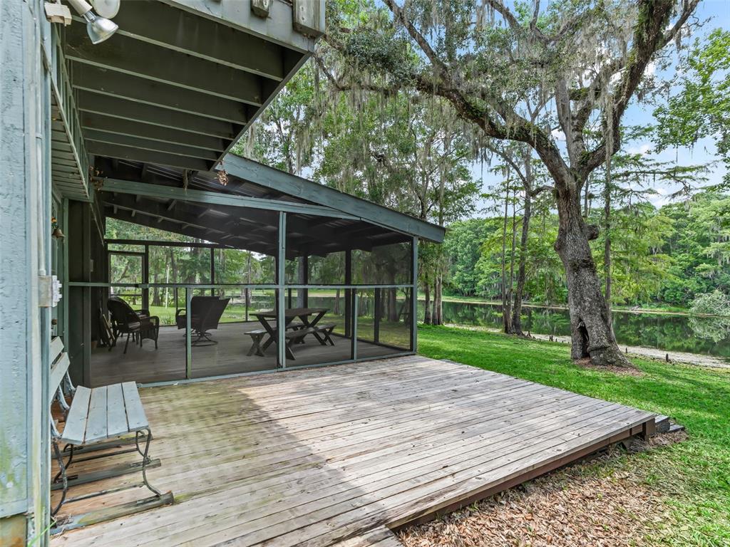 A wooden deck leads to a screened-in porch overlooking a lake with large trees — 10518 SW 64TH AVENUE, BUSHNELL, FL