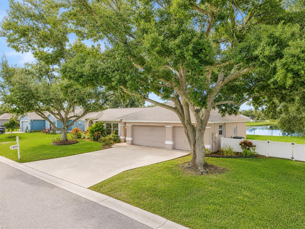 A single-story house with a two-car garage and large trees sits on a grassy lot — 6305 61ST STREET E, PALMETTO, FL