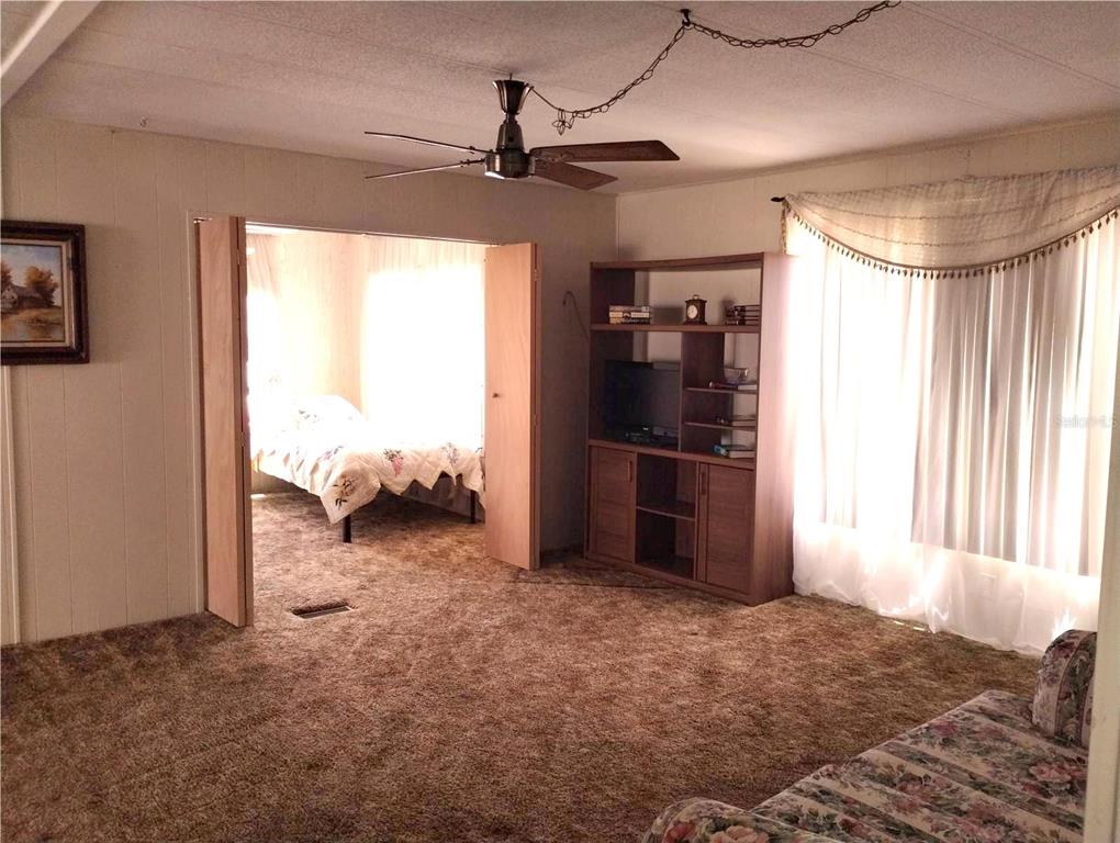 A carpeted living room features a ceiling fan, a wooden shelving unit, and an adjacent bedroom — 7409 SKYLINE DRIVE, HUDSON, FL