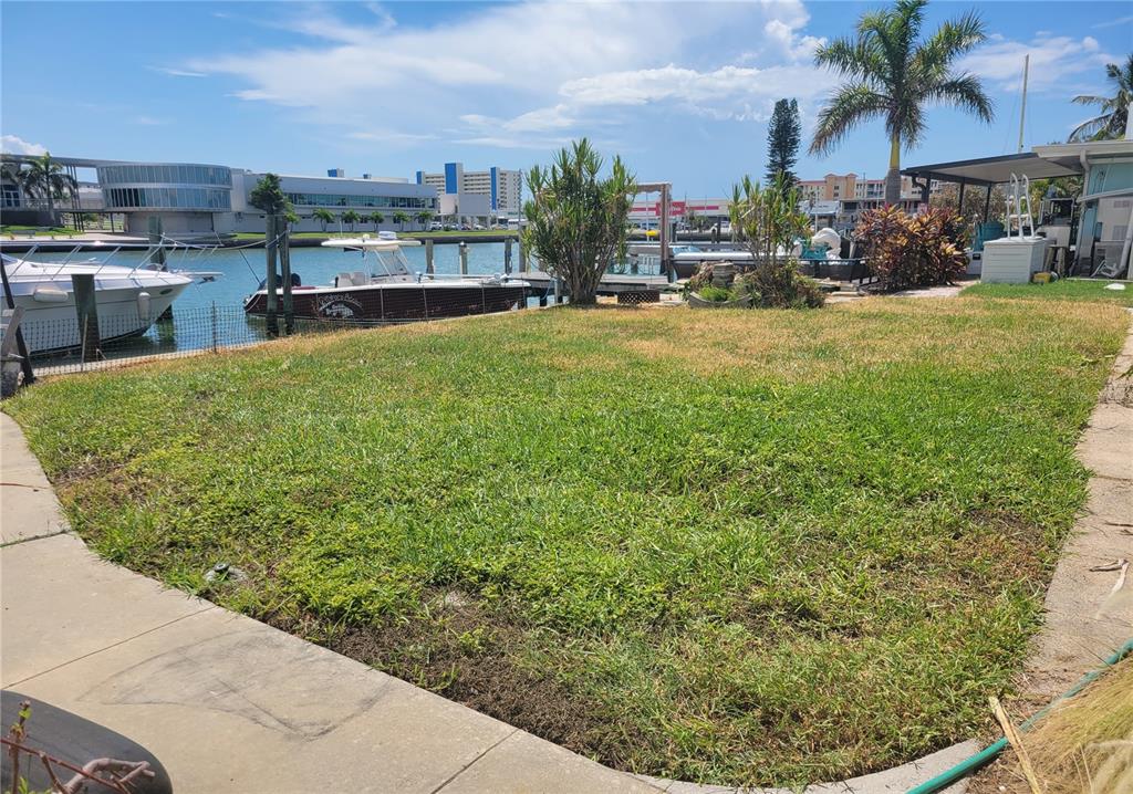 A grassy backyard overlooks a canal with docked boats and nearby waterfront buildings — 15318 HARBOR DRIVE, MADEIRA BEACH, FL