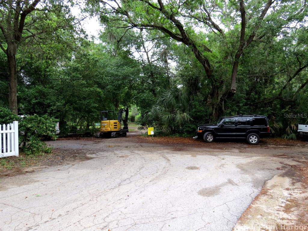 A yellow excavator sits on a dirt path next to a black SUV among trees — ROBERTS ROAD, PALM HARBOR, FL