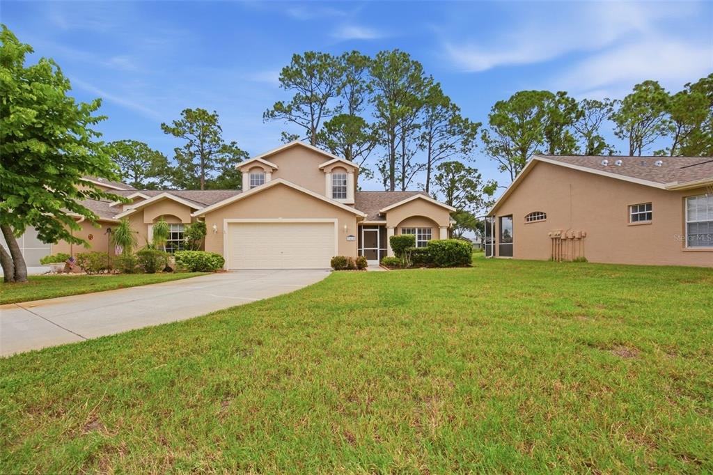 A tan suburban house with a two-car garage, a front lawn, and surrounding pine trees — 7444 MOORGATE COURT, NEW PORT RICHEY, FL
