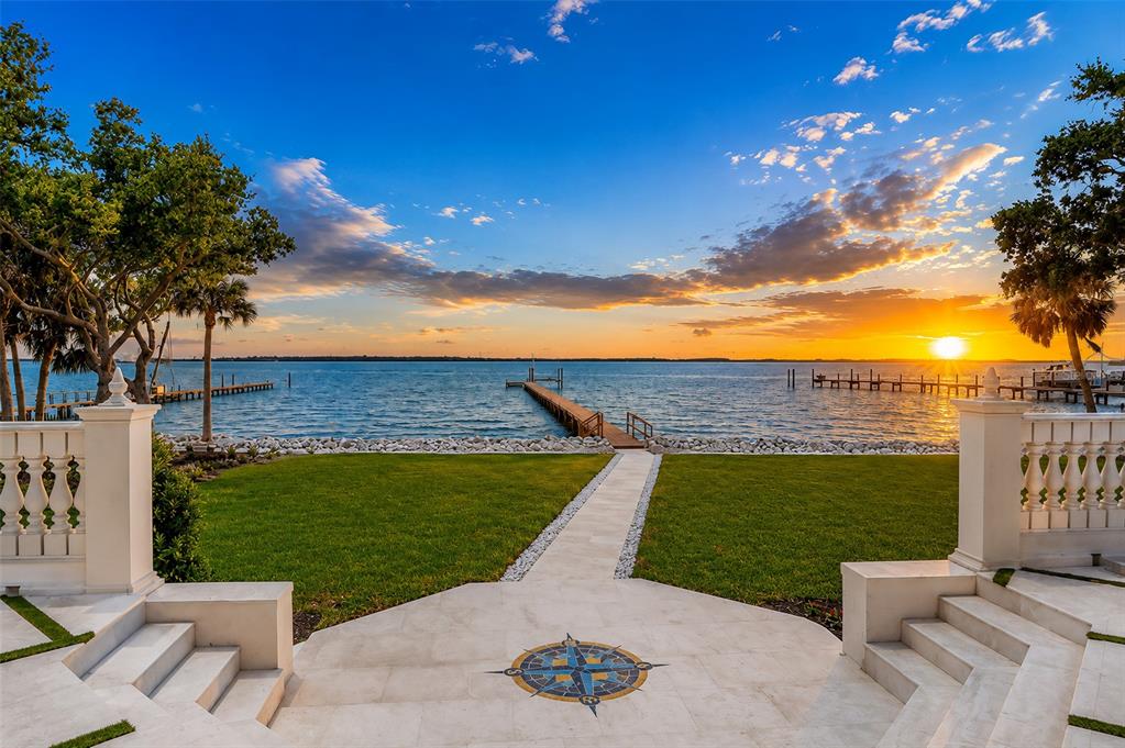 A stone patio with a compass inlay leads to a lawn, dock, and sunset over water — 1516 BAYSHORE BOULEVARD, DUNEDIN, FL