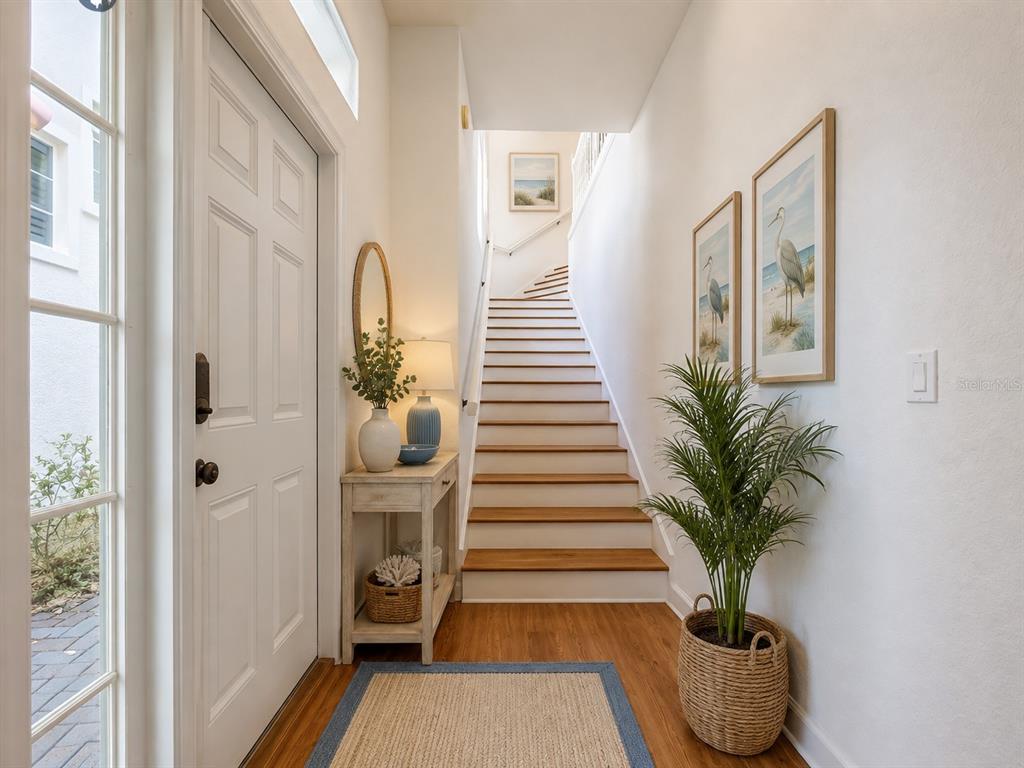 A bright entryway features a white door, a wooden staircase, and coastal-themed wall art — 1338 EMERALD DUNES DRIVE UNIT 35, SUN CITY CENTER, FL