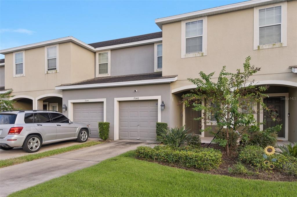 A two-story townhouse with a gray garage door, a concrete driveway, and a small front garden — 7046 TOWNE LAKE ROAD, RIVERVIEW, FL