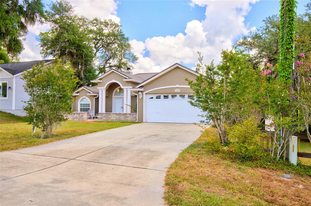 A tan single-story house with a white garage door and arched entryway sits behind a driveway — 13059 NE 7TH LOOP, SILVER SPRINGS, FL
