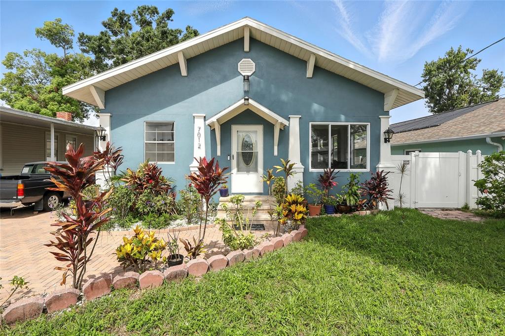 A blue bungalow with a white front door, brick walkway, and tropical landscaping — 7012 N HUBERT AVENUE, TAMPA, FL