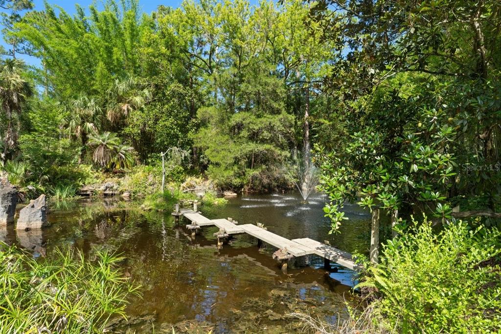 A wooden footbridge crosses a pond with a fountain in a lush, wooded backyard setting — 400 PINE BLUFF TRAIL, ORMOND BEACH, FL