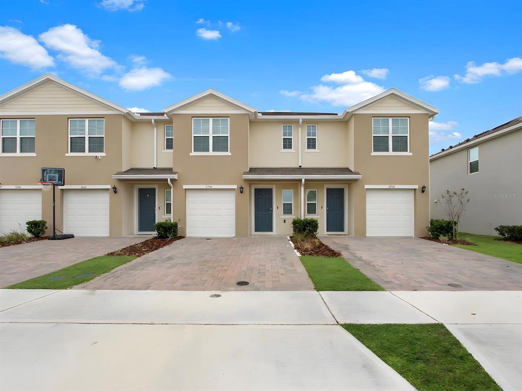 A row of four attached townhomes with tan stucco exteriors, white garage doors, and paved driveways — 2798 BRIE HAMMOCK BEND, HARMONY, FL