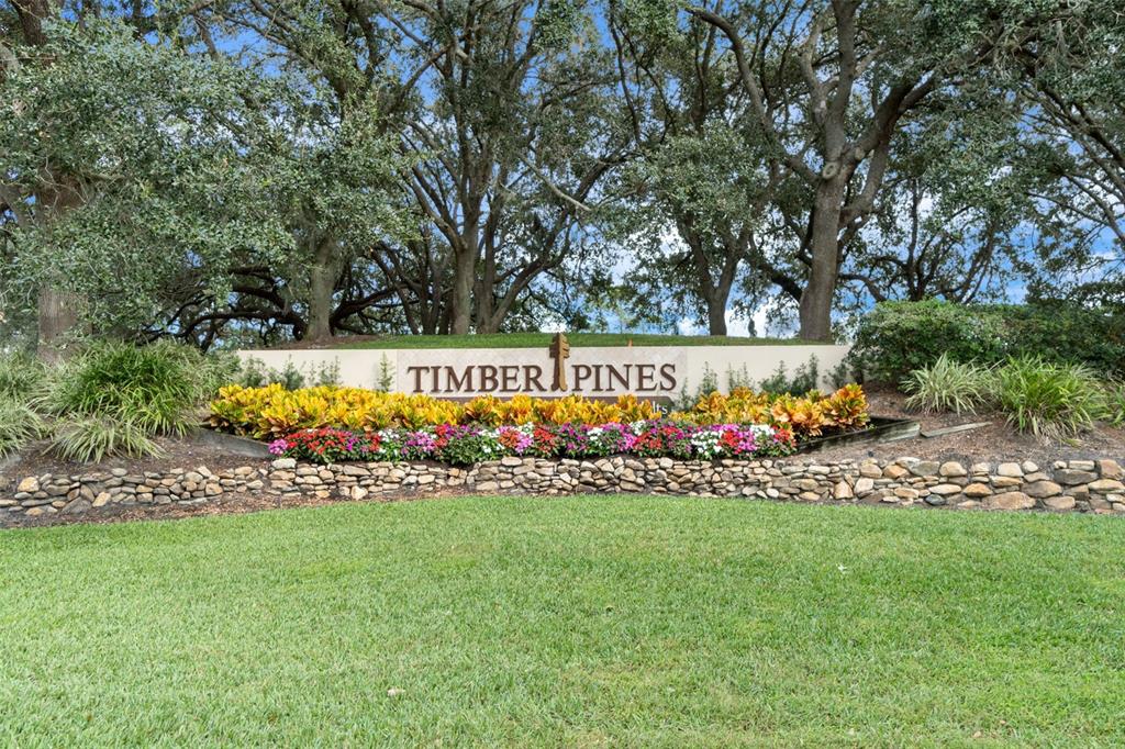A stone retaining wall sits in front of a Timber Pines community entrance sign — 7196 CROWN OAKS DRIVE, SPRING HILL, FL