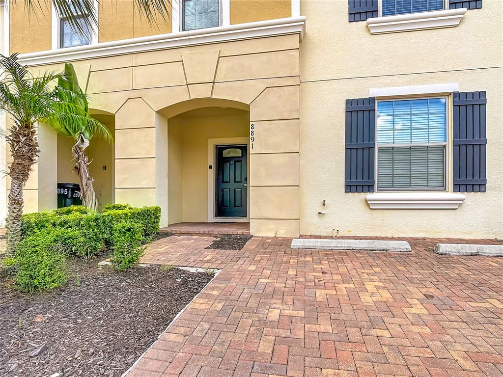 A tan townhouse exterior features a dark front door, brick paver driveway, and small palm tree — 8891 GENEVE COURT, KISSIMMEE, FL