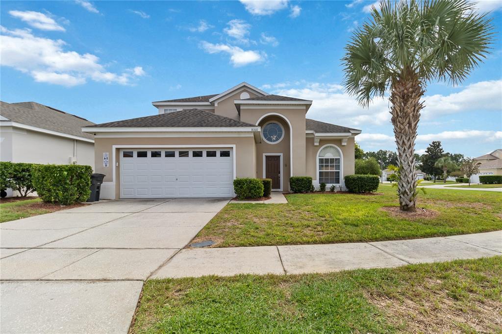 A tan two-story house features a white garage door, a palm tree, and a sidewalk — 8160 SUN PALM DRIVE, KISSIMMEE, FL
