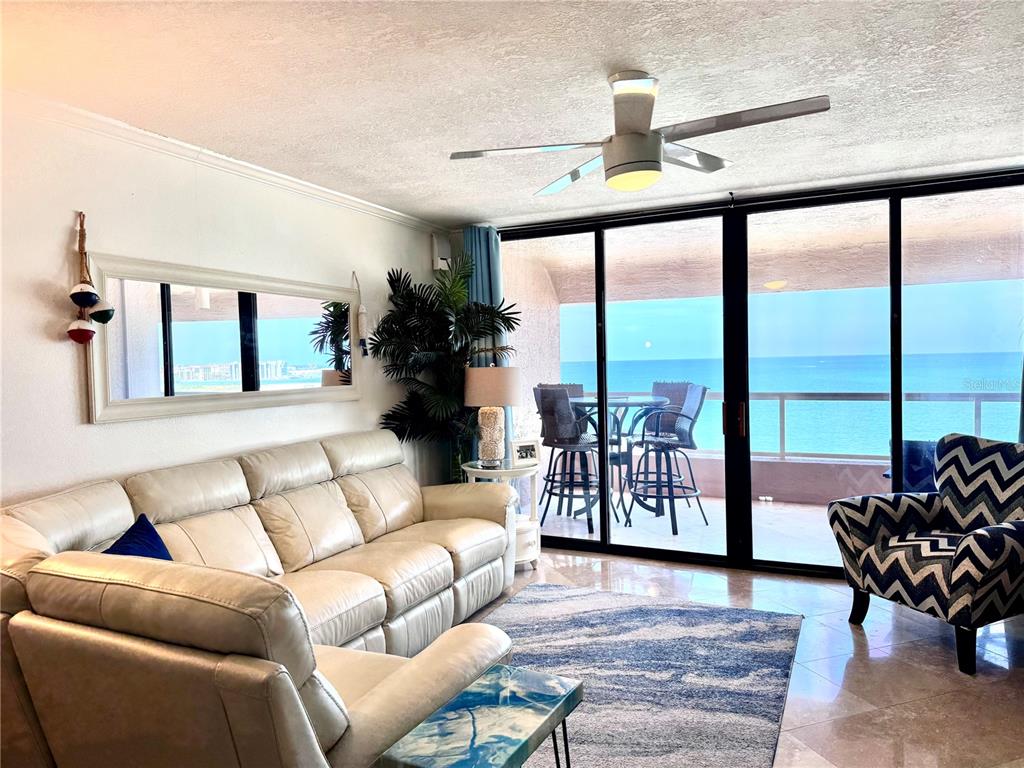 A living room features a white leather sofa, patterned armchair, and sliding doors overlooking the ocean — 1310 GULF BOULEVARD UNIT 18B, CLEARWATER, FL