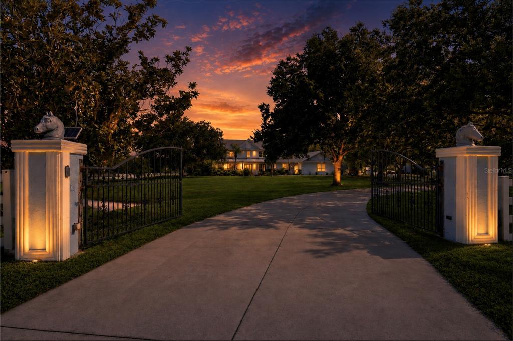 A concrete driveway leads through gated stone pillars toward a house at sunset — 20207 77TH AVENUE E, BRADENTON, FL