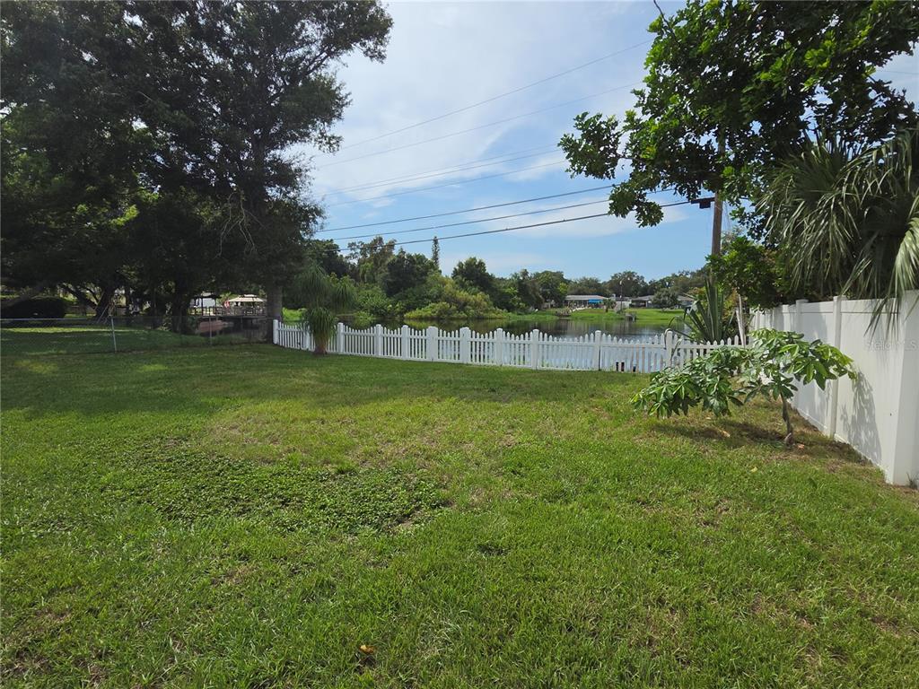 A grassy backyard features a white picket fence overlooking a small pond with trees — 10827 VILLAGE GREEN AVENUE, SEMINOLE, FL