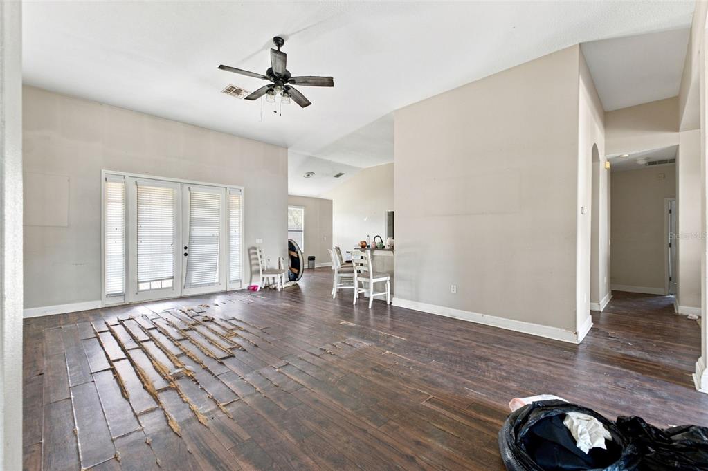 A living room with damaged wood flooring, a ceiling fan, and a view into the kitchen — 5598 THORNGROVE WAY, SPRING HILL, FL