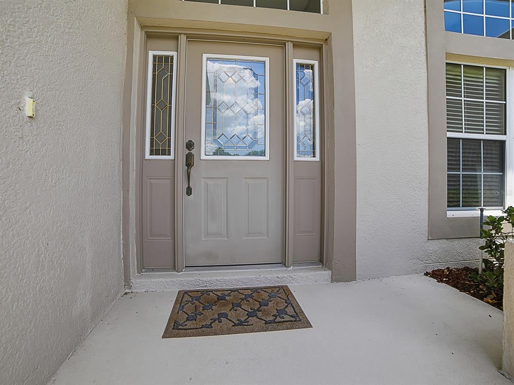 A beige front door with glass inserts and sidelights sits on a concrete porch — 5106 CHANTICLEER DRIVE, LEESBURG, FL
