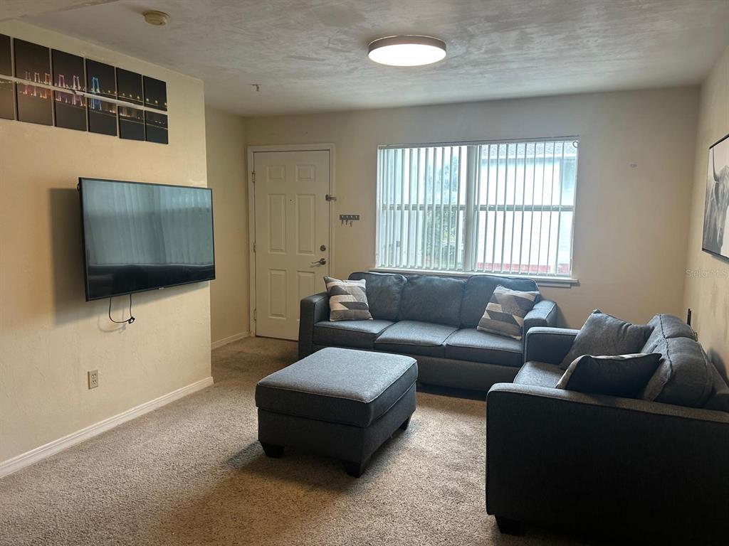 A living room features a wall-mounted television, gray sofa, matching ottoman, and vertical window blinds — 2360 SW ARCHER ROAD UNIT 915, GAINESVILLE, FL