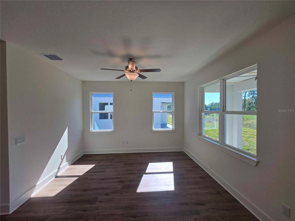 An empty room features wood-look flooring, a ceiling fan, and multiple windows with outdoor views — 3352 DAYBREAK RUN LOOP, SPRING HILL, FL
