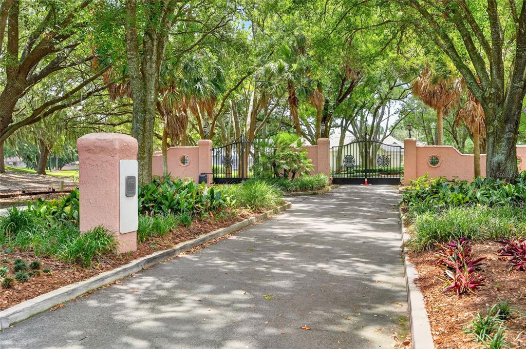 A paved driveway leads to a gated entrance flanked by pink stucco walls and trees — 5 HICKORY HEAD HAMMOCK, THE VILLAGES, FL