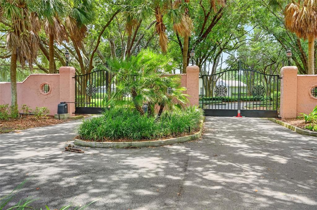 A gated entrance with pink stucco walls and lush landscaping leads to a paved driveway — 5 HICKORY HEAD HAMMOCK, THE VILLAGES, FL