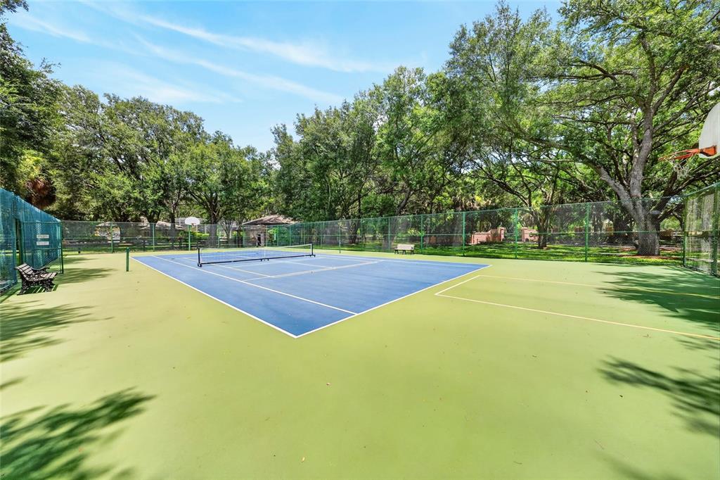 A fenced outdoor tennis court with a blue surface surrounded by green trees — 5 HICKORY HEAD HAMMOCK, THE VILLAGES, FL