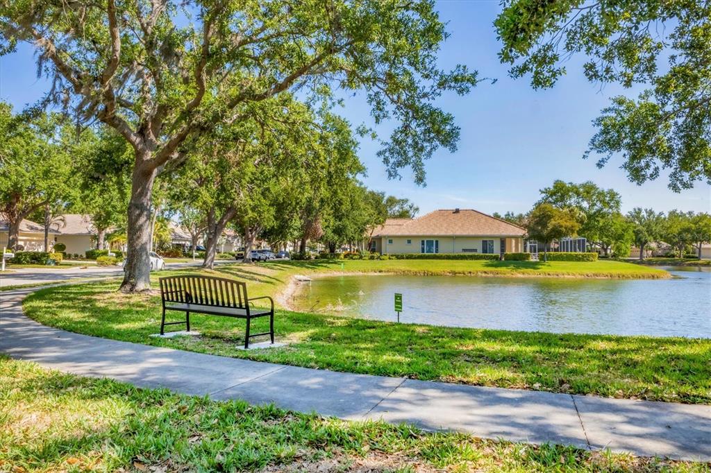 A park bench sits on a grassy lawn beside a pond near a residential street — 4358 MIRABELLA CIRCLE, BRADENTON, FL