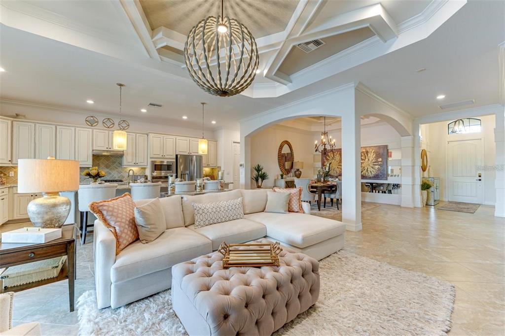 A bright, open-concept living room features a tufted ottoman, sectional sofa, and kitchen beyond — 5803 CESSNA RUN, BRADENTON, FL