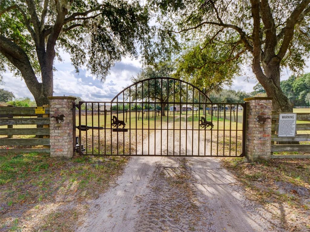 A metal arched gate with horse silhouettes stands between two brick pillars on a dirt road — 1765 E CR 462, WILDWOOD, FL