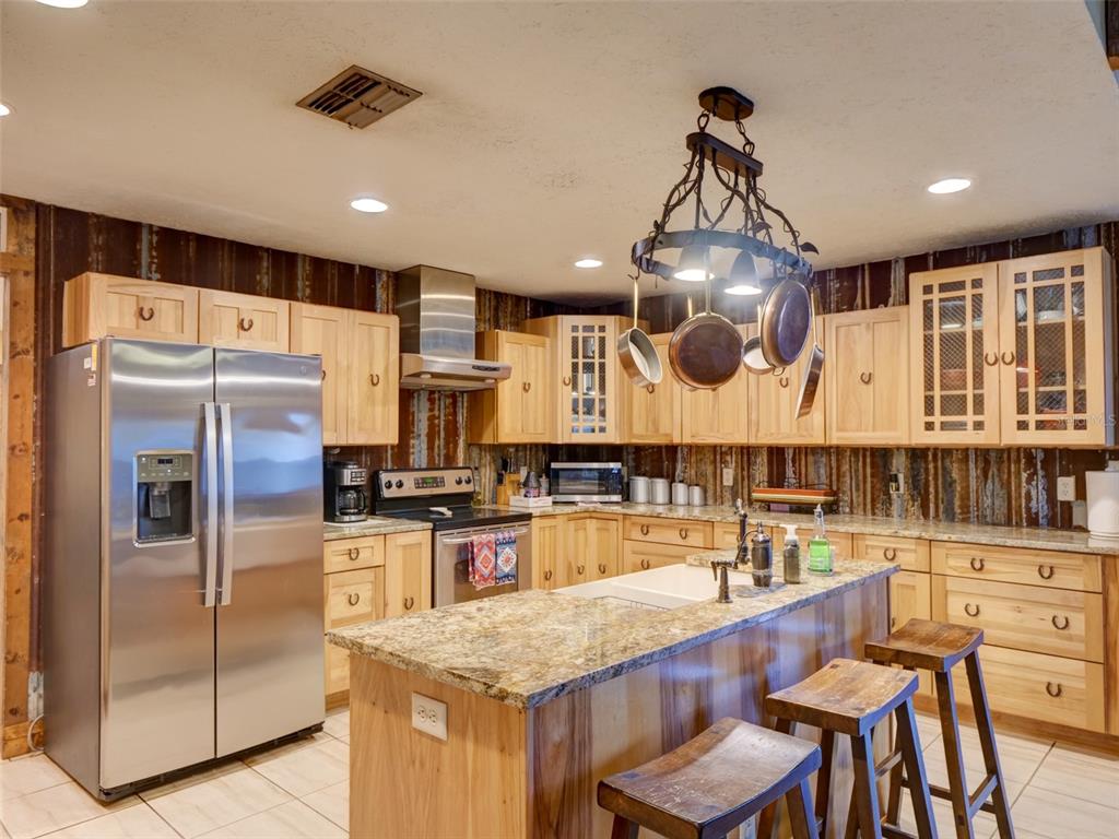 A kitchen features light wood cabinets, granite countertops, a central island, and a rustic pot rack — 1765 E CR 462, WILDWOOD, FL